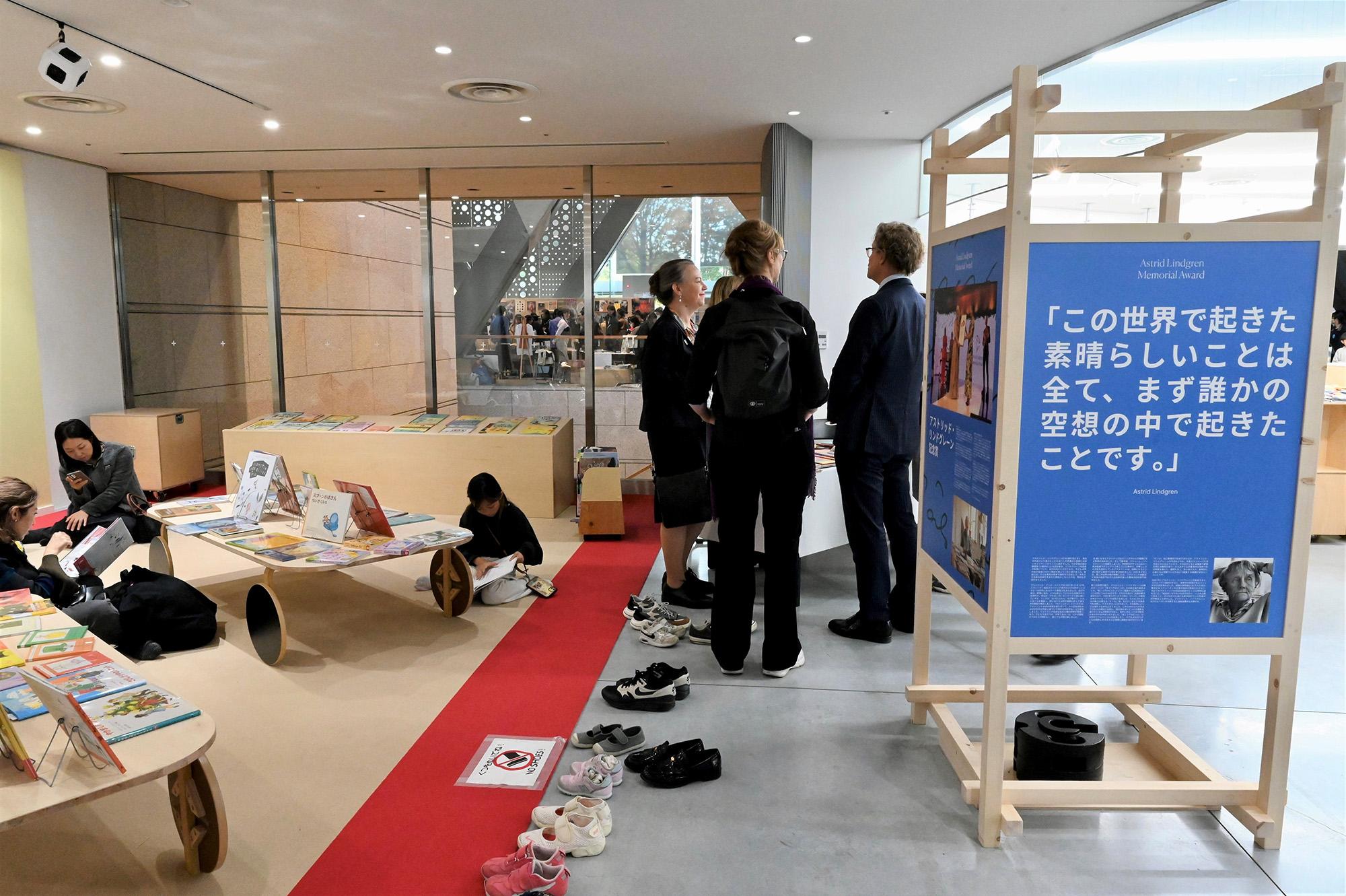 Children and adults reading books, shoes parked outside the reading area. 