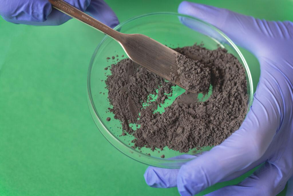 Gloved hands holding a lab dish with a grey powder in it.