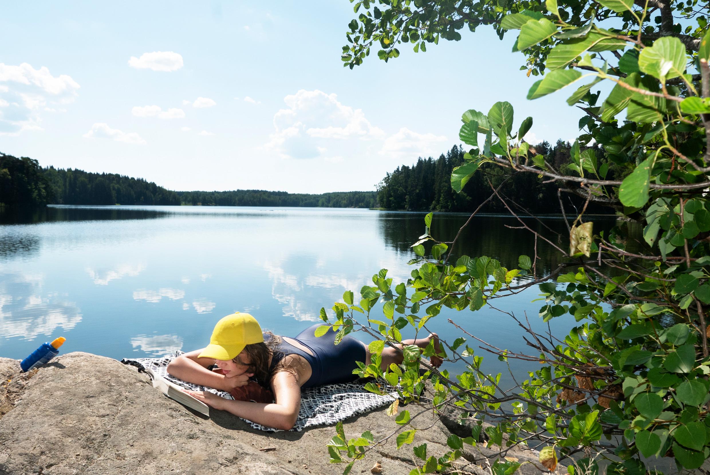 A girl in a yellow cap, reading a book on a cliff by a blue lake.