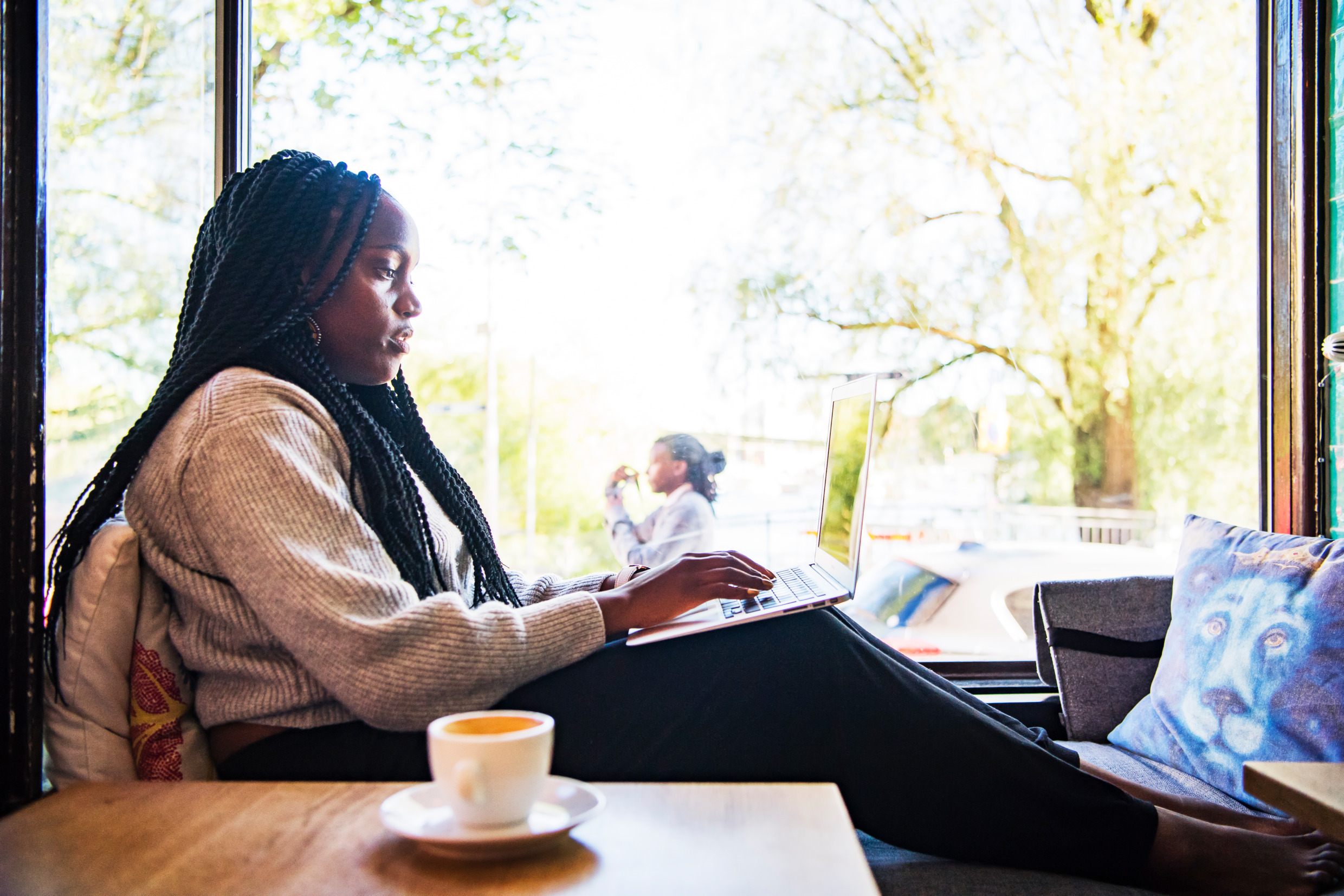 A woman is sitting inside by a window with a cup of coffee, typing on a laptop in her lap.